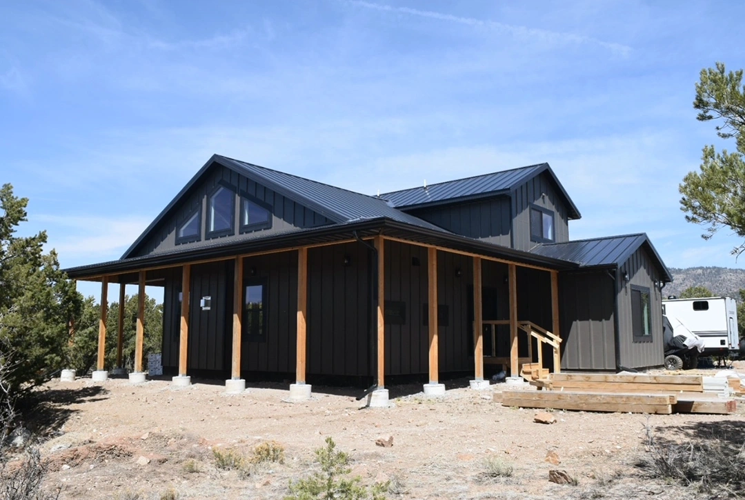 Exterior view of a completed custom home featuring dark metal siding with light trim accents, metal roofing, wraparound covered porch with wooden support posts, and mountain terrain landscaping under a blue sky with white clouds