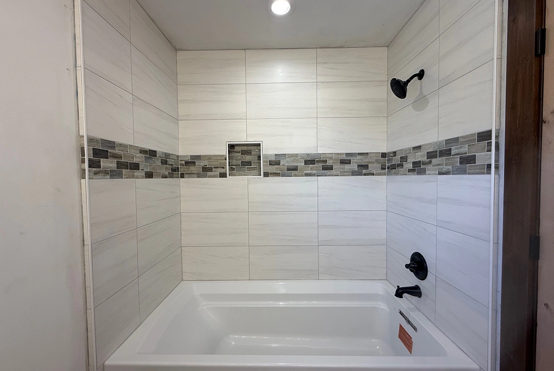 Custom home bathroom tub and shower combination with large-format white tile walls, a decorative grey mosaic tile accent band, built-in shower niche, and matte black fixtures