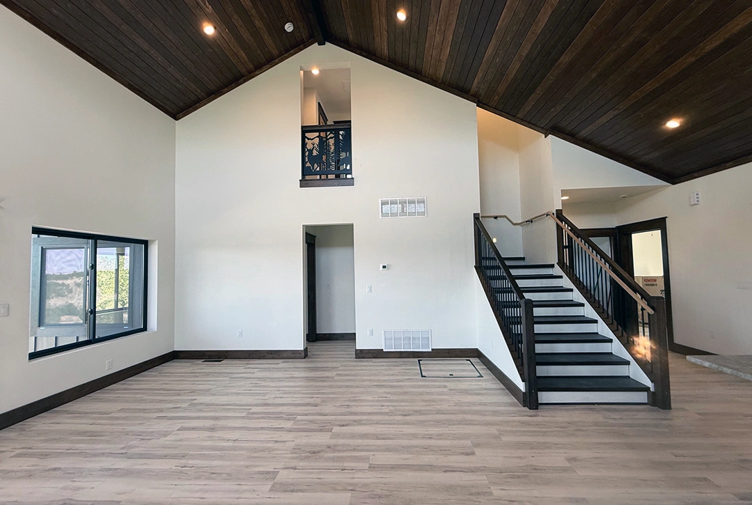 Custom home interior with soaring vaulted ceiling in dark stained tongue-and-groove wood, white walls, open staircase with black railings and a custom pine-tree metal panel accent, and a loft overlook