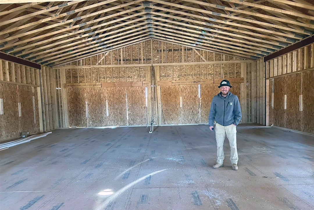Interior of a large framed building showing Derek True standing in the center of an open space with exposed wooden ceiling trusses, OSB wall sheathing, and a concrete floor with visible radiant heating lines